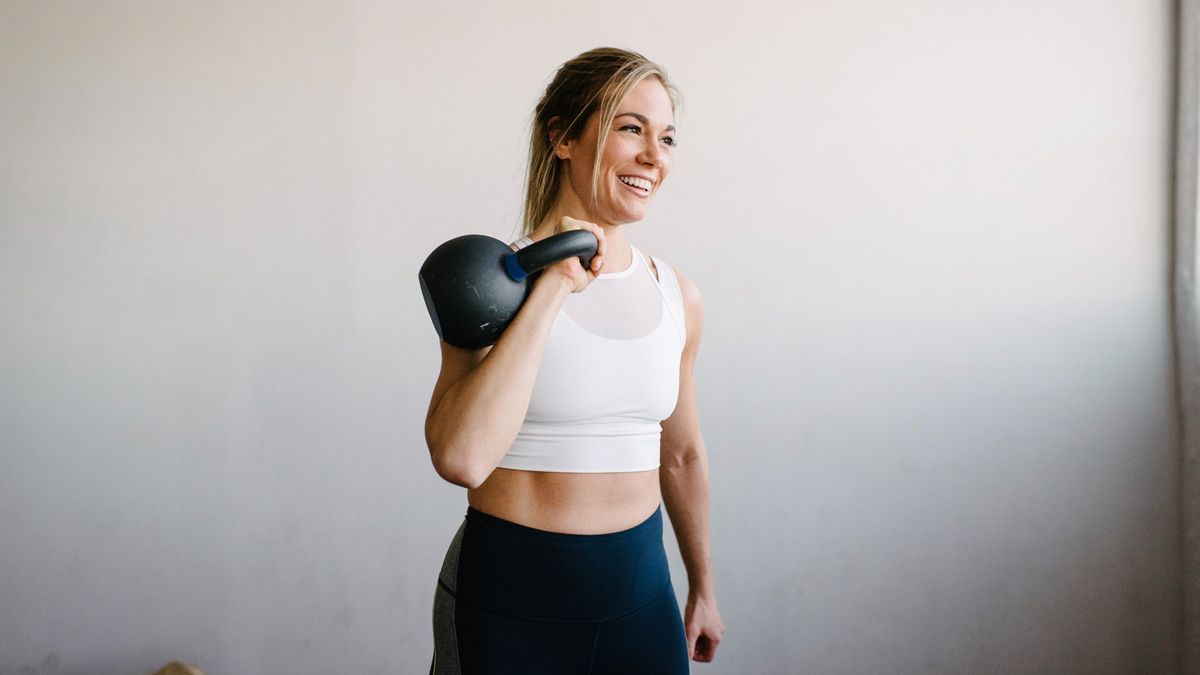 Woman holding a kettlebell in a racked position against her shoulder, smiling, against a white back drop