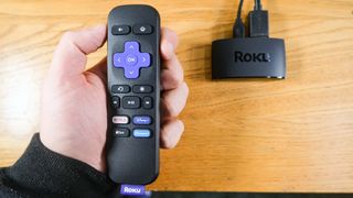 A hand holds the remote for the Roku Express (2022) above the streaming device
