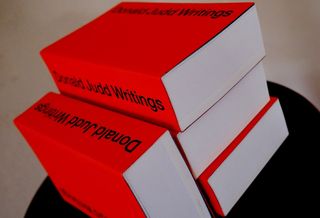 Four books with a red cover say 'Donald Judd Writings'. They're photographed on a black stool.