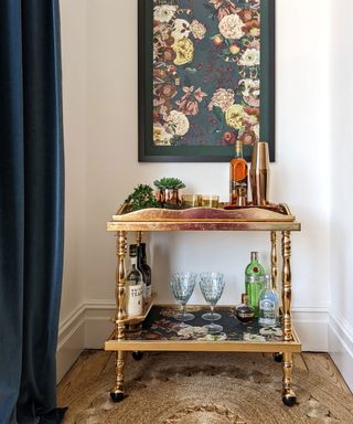 Upcycled gold drinks trolley, decorated with drinks and glassware, wooden flooring, woven rug, artwork on wall mounted above, dark blue curtains