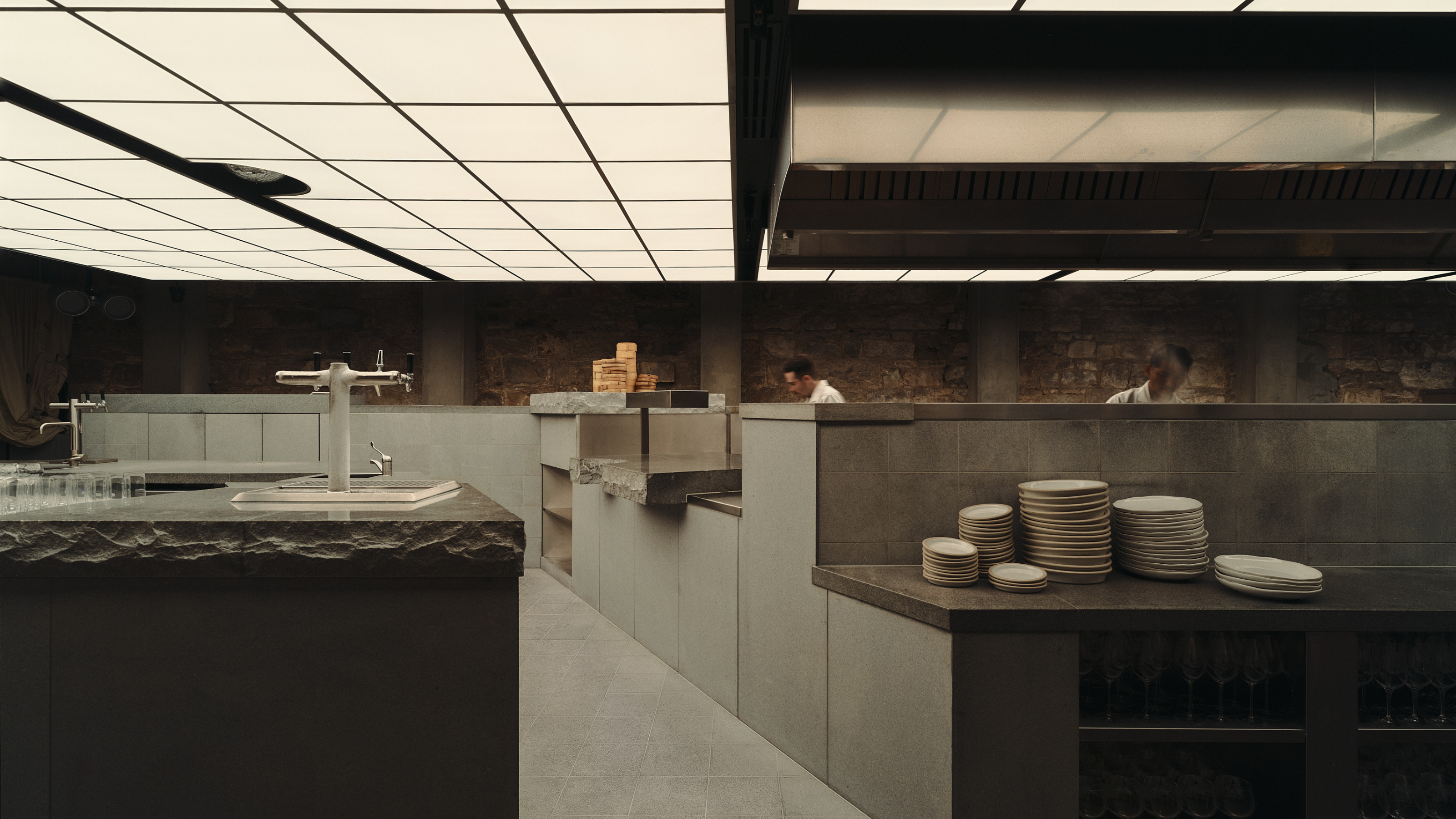 a restaurant in a brutalist design style with stone counter tops, a ceiling covered in ceiling lights, and a stack of several plates on the counter