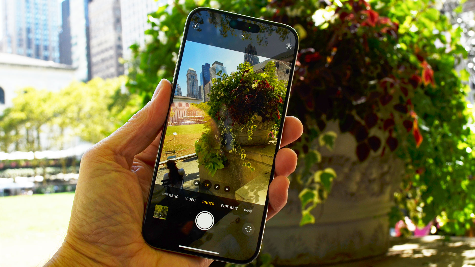 Apple iPhone 15 Pro Max i hånden med kameraet tændt og en blomst i baggrunden.