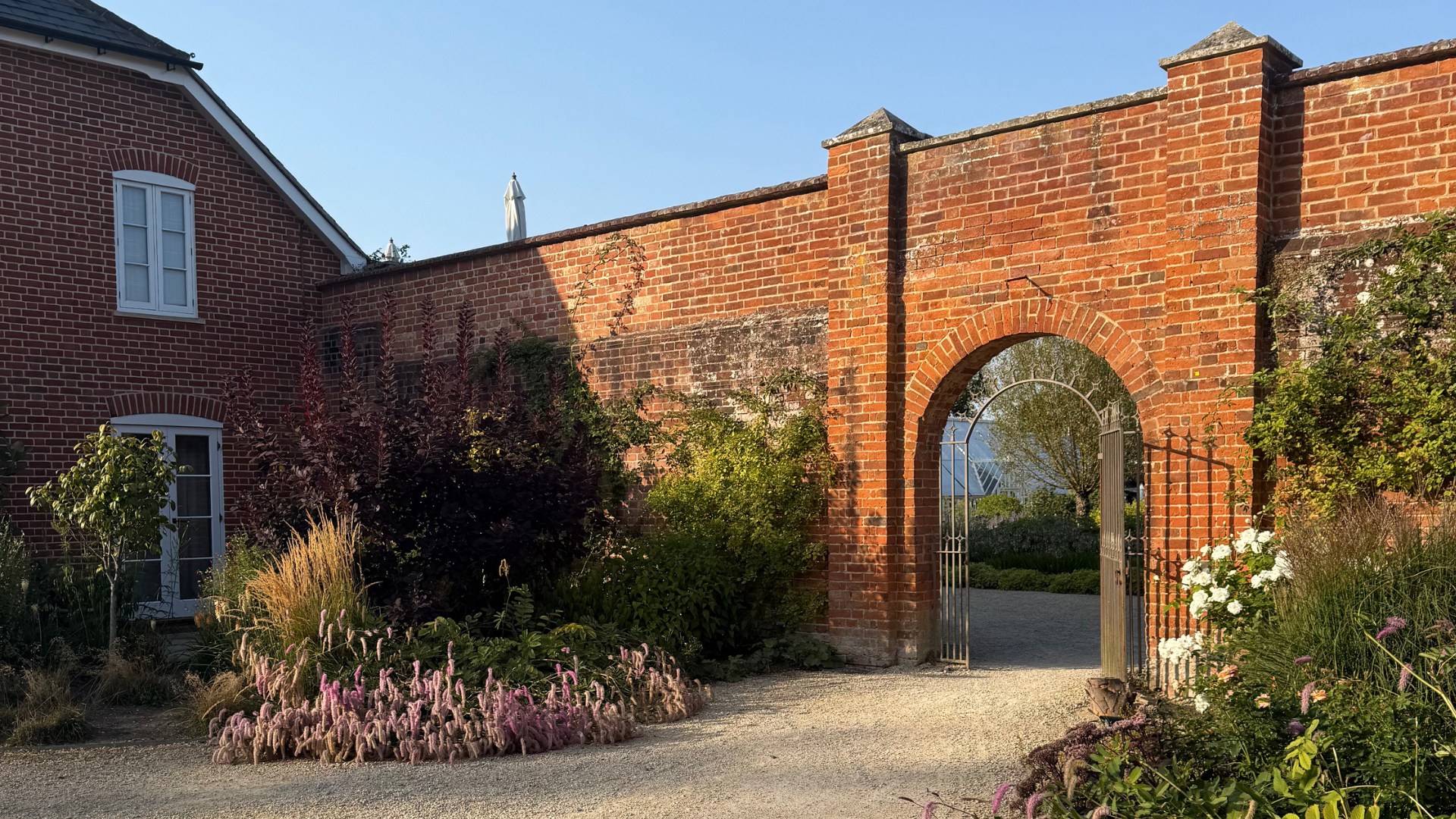 A sunlit walled garden with soft pink and purple flowers spilling onto a gravel path. A red brick wall with an arched iron gate frames the view beyond, next to a traditional brick building under a clear blue sky.