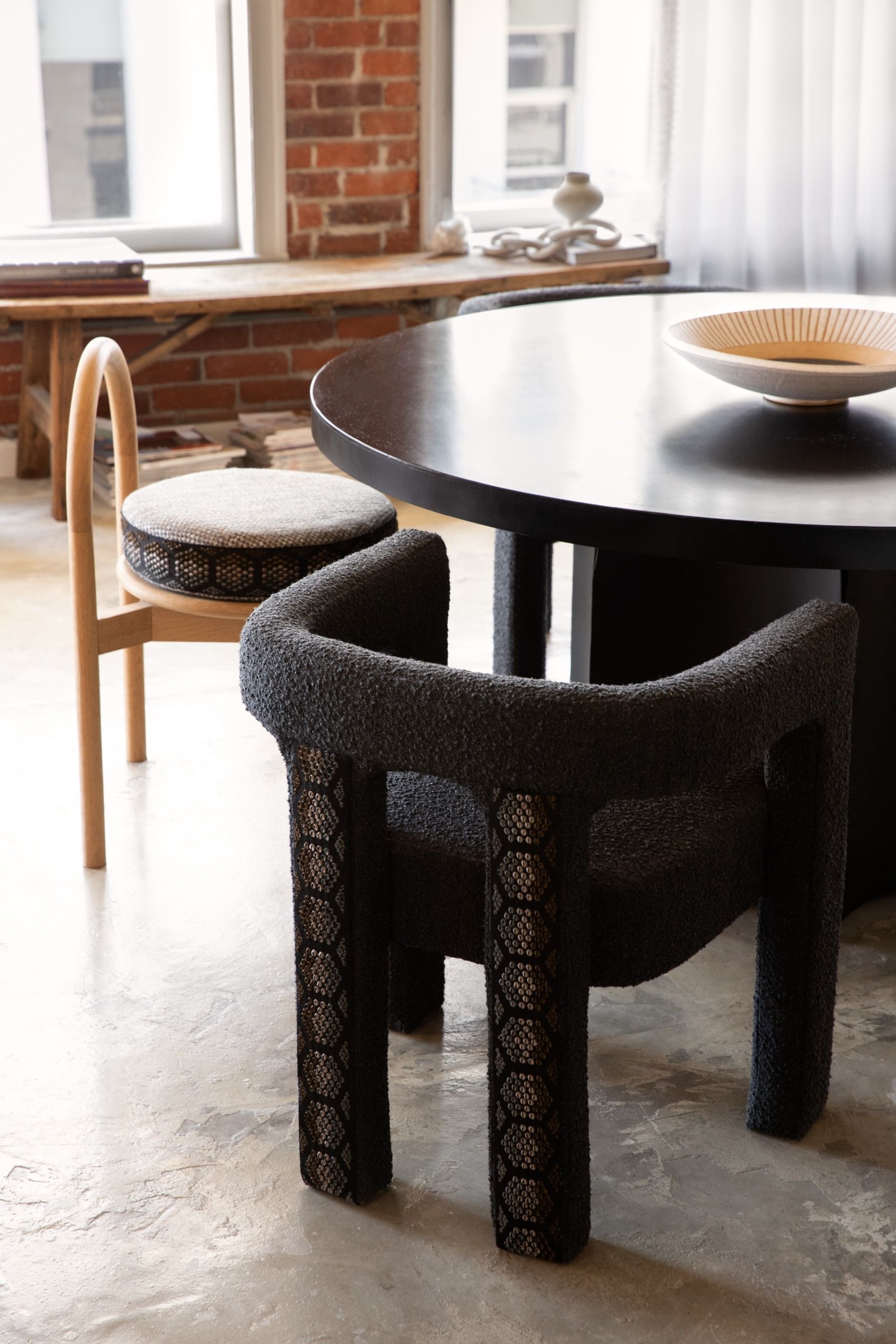Loft-style apartment with exposed brick wall, low-profile wooden bench, circular black table, black boucle dining chair edged with studs in a hexagonal pattern siting on poured concrete flooring
