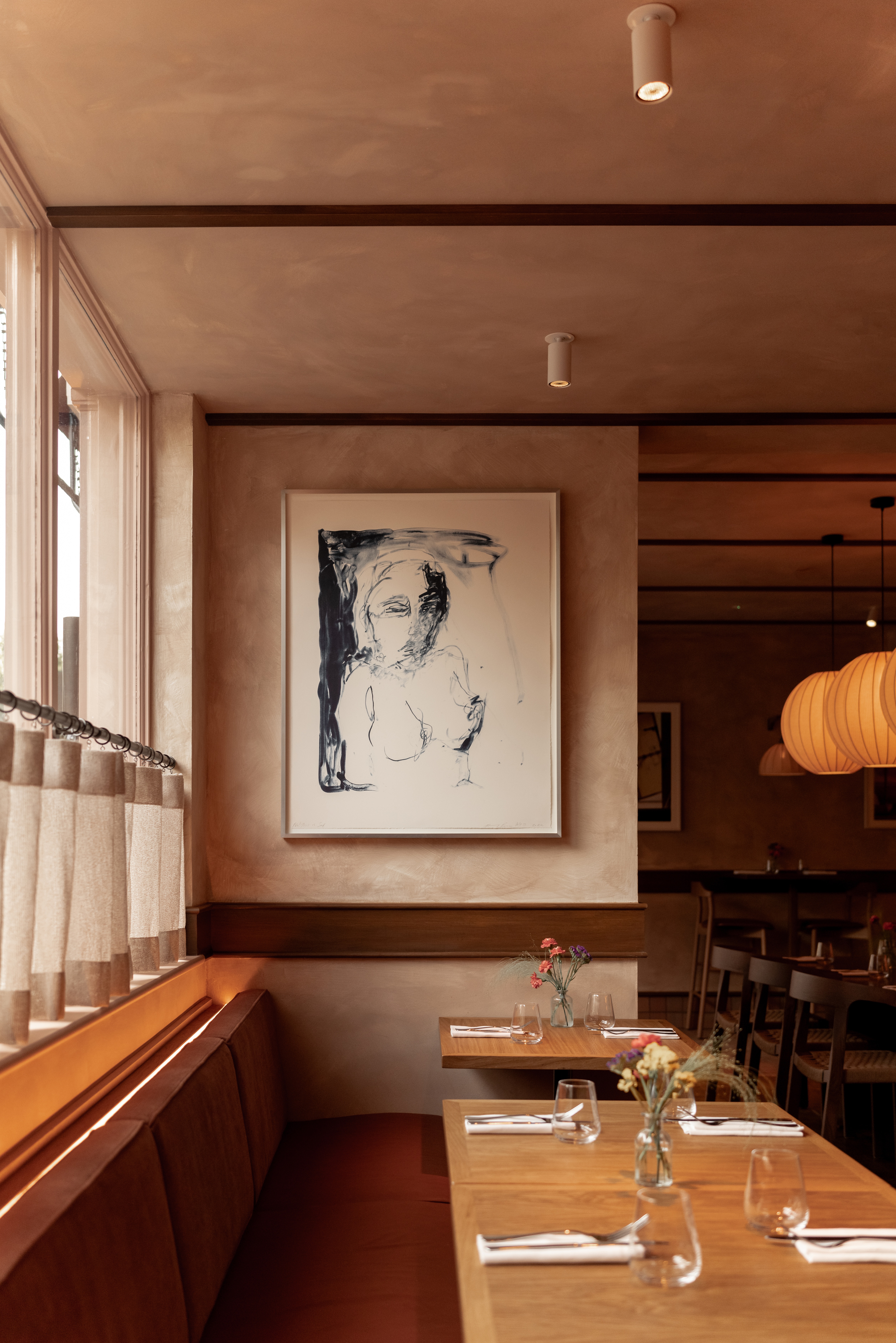 A soulful, retro-chic restaurant is decorated with cafe style curtains, wood and velvet furnishings, original artwork, and warm, lantern-shaped pendant lights in paper.