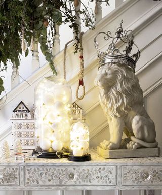 White porcelain lion with silver crown next to glass cloche with white fairy lights next to white staircase decorated with green foliage