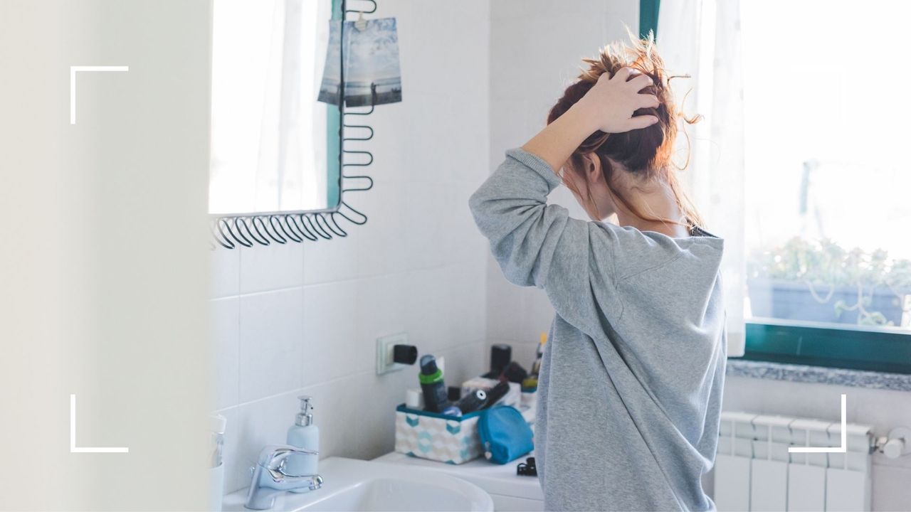 Woman holding her sore scalp in the bathroom, looking in the mirror with back to the camera next to large open window