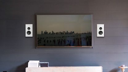 PSB PWM Sat speakers in white on a dark wall, either side of a wall-mounted TV