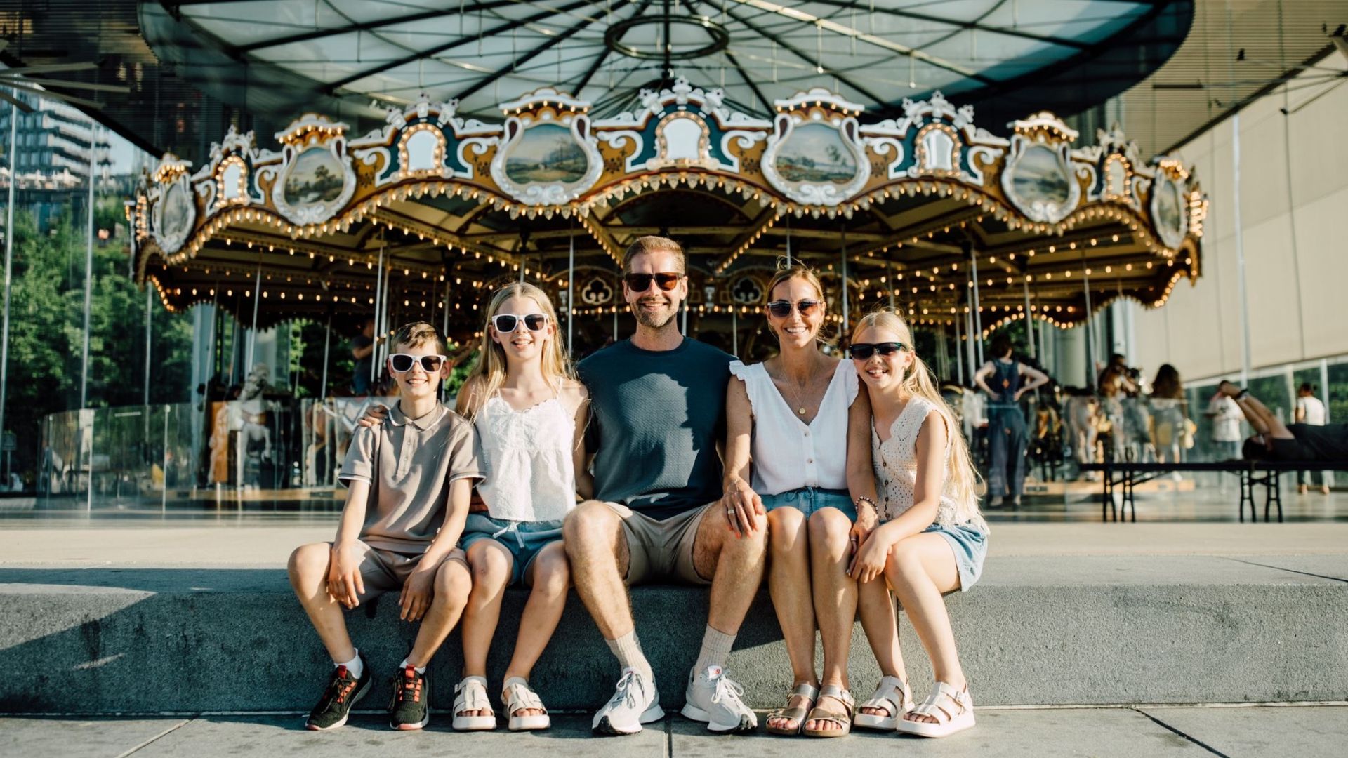 Laura Dove with her husband and three of her children sit in front of a carousel at a funfair, all wearing shorts, t-shirts and sunglasses