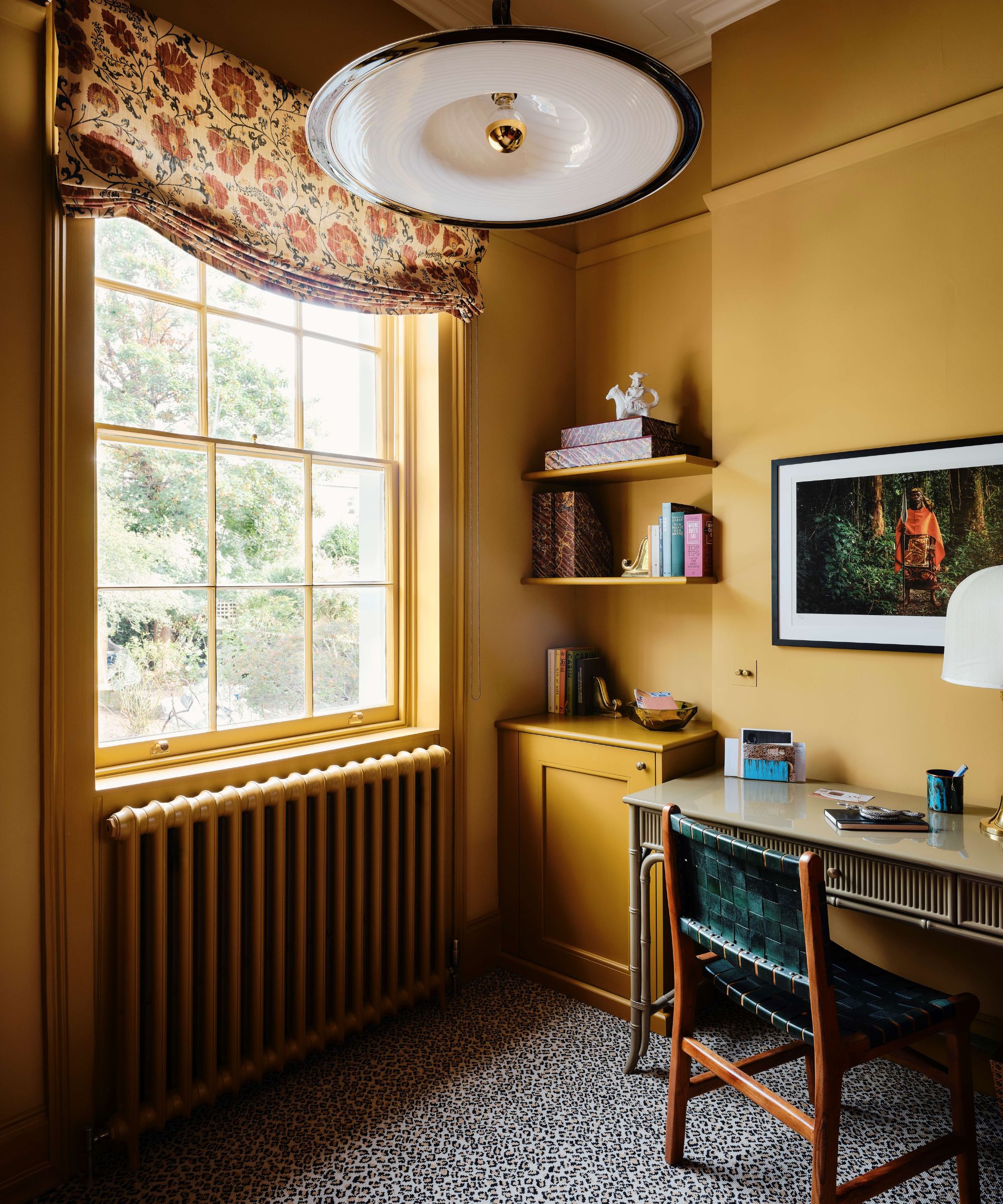 A home office with ochre color-drenched walls, a window with a floral roman blind, a column radiator, and a desk with a teal chair.