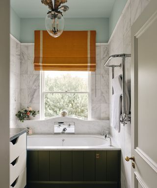 neutral bathroom with tiled marble walls and a green panelled bath, pale blue ceiling and an orange window blind