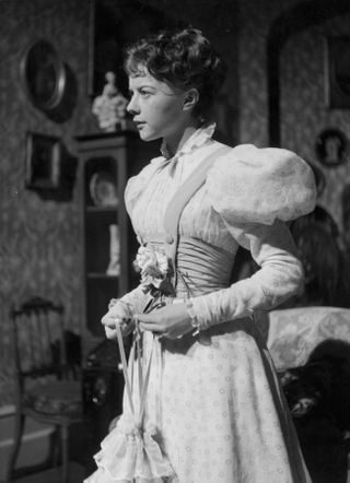 a woman wearing a gown with puffy sleeves stands in a living room in the original version of the importance of being earnest