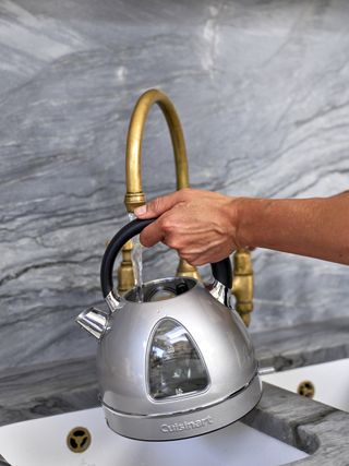 hand filled silver metallic kettle from swan neck brass tap in kitchen with gray marble backsplash