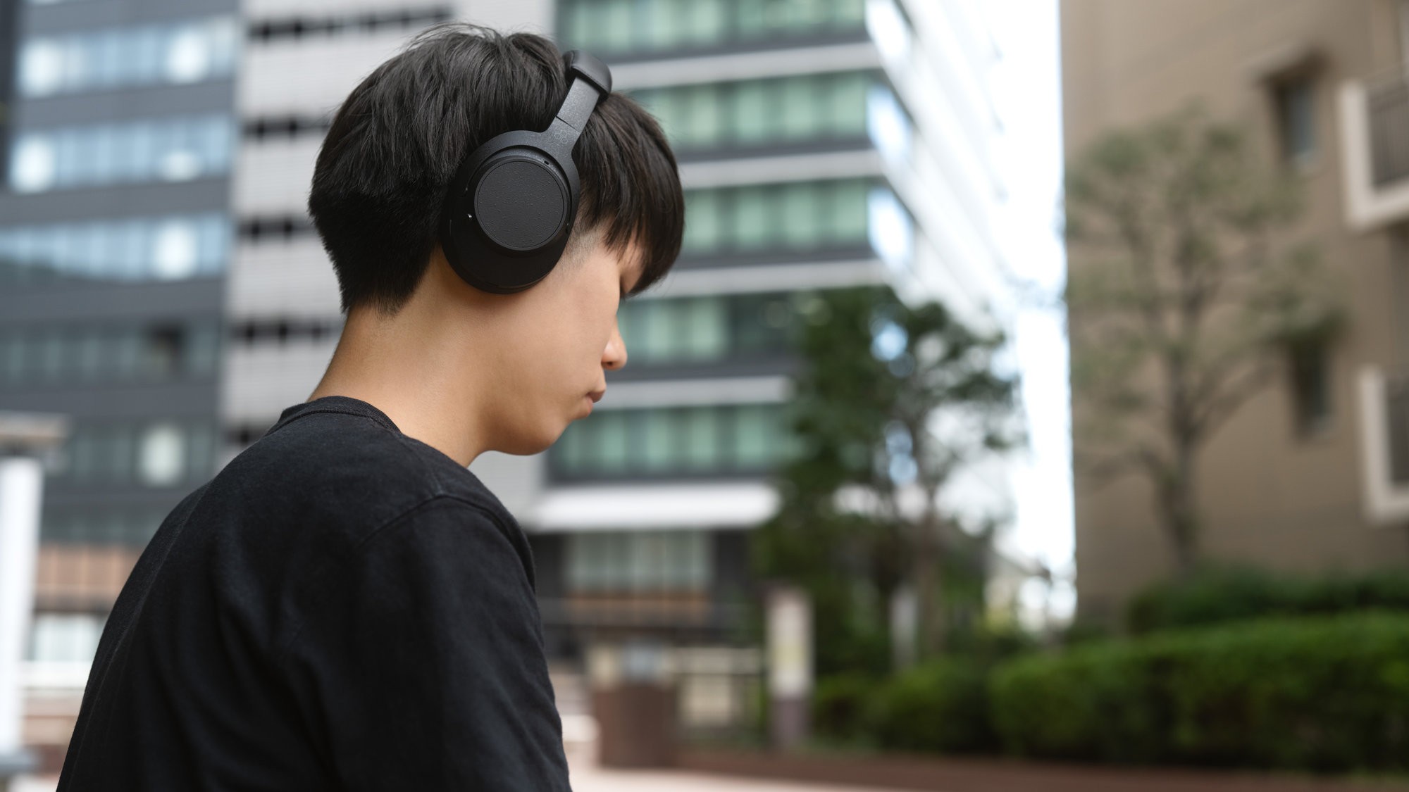 A young person wearing a black t-shirt has their back to the camera, wearing black over-ear headphones (likely a Final UX5000) while standing outside an office or apartment building.