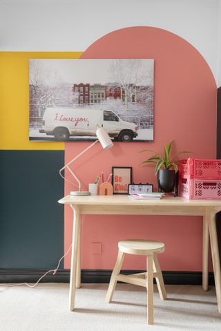 Home office area with oak desk and stool, angled white desk lamp, and colour-blocked walls made up of green and yellow squares and a pink arch