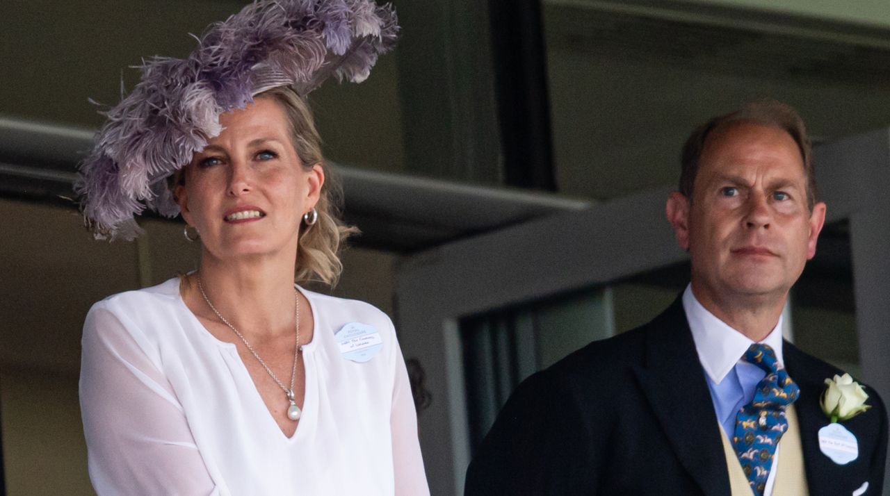 Duchess Sophie wears a sheer white shirt and large purple hat featuring lots of feathers, and husband Prince Edward stands beside her