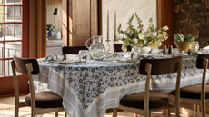 a dining table with a blue floral tablecloth, places set with plates, napkins, utensils, glassware, and a table decorated with a pitcher, candle holders, a floral display, a decorative bowl, and salt and pepper shakers. Brown dining chairs surround the table