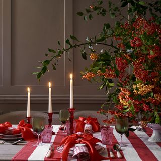 A dining table covered in a striped runner with a large red berry centrepiece and napkins on the plates held together with red satin bows