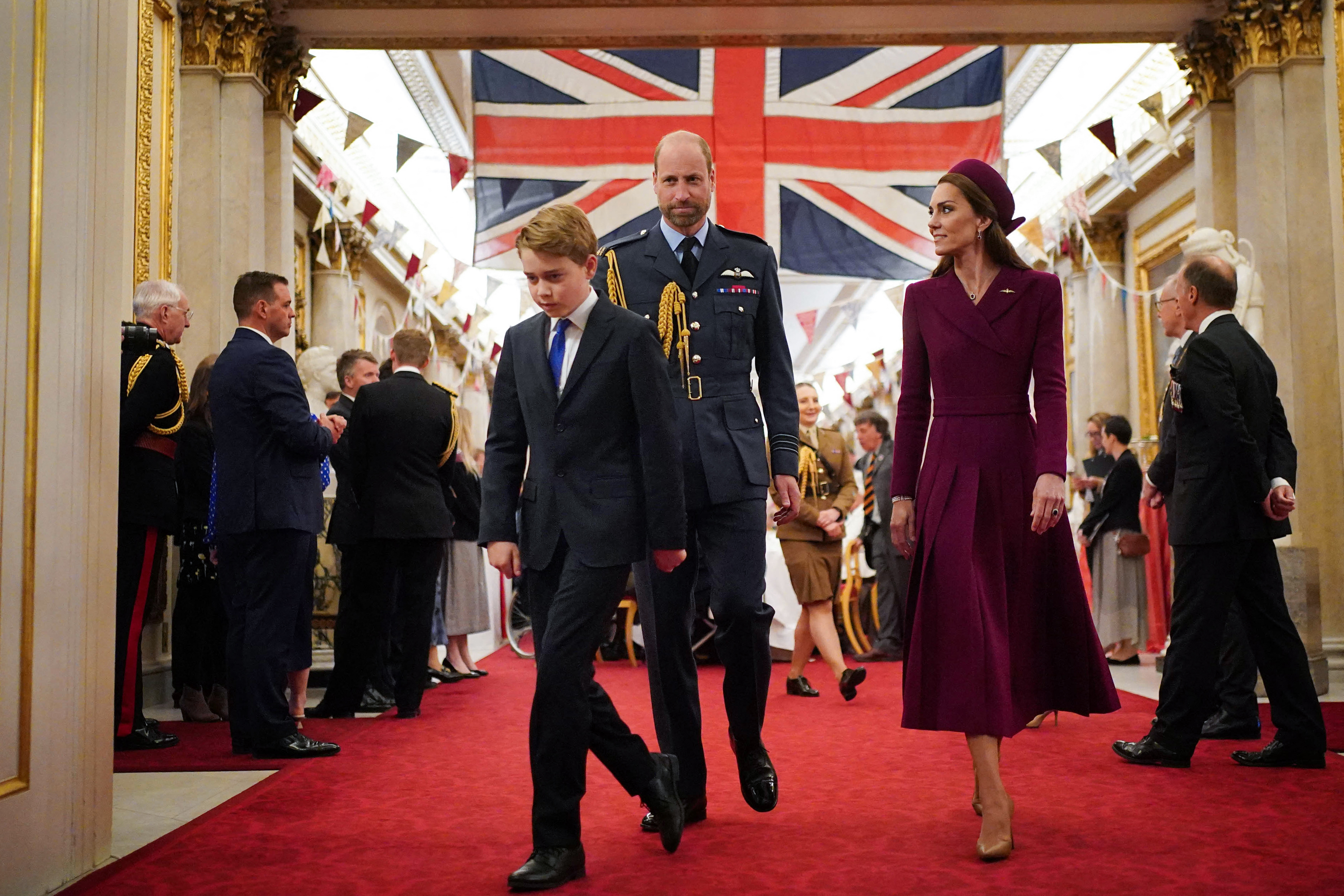 Prince George walking in front of Prince William and Princess Kate on a red carpet with a huge Union Jack above them