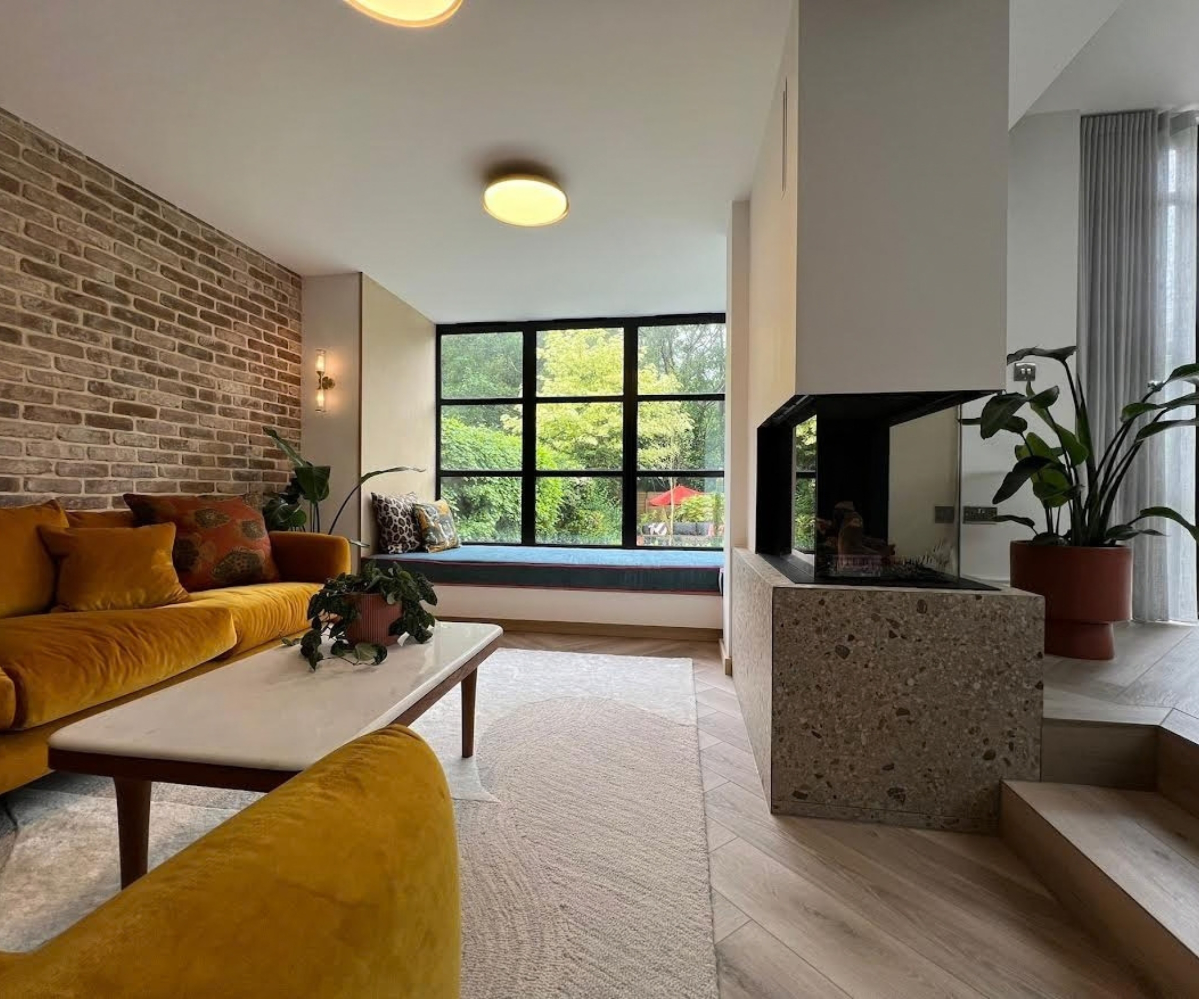 Living room with mustard velvet sofa, exposed brick wall, terrazzo fireplace, and a garden window seat.