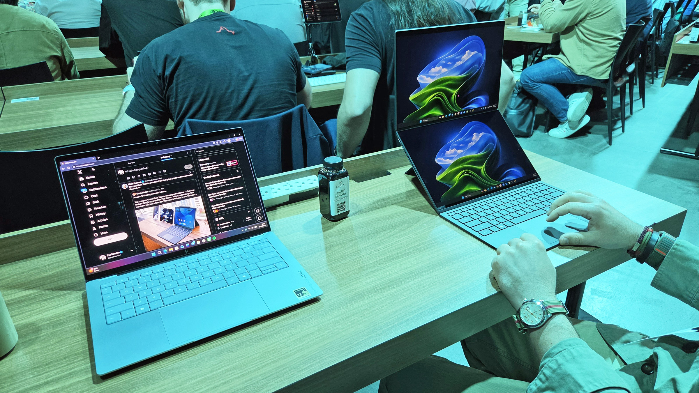 A person uses the detached keyboard of an ASUS Zenbook Duo laptop configured in a stacked vertical dual-screen setup on a wooden desk, sitting next to another open laptop at a tech event.