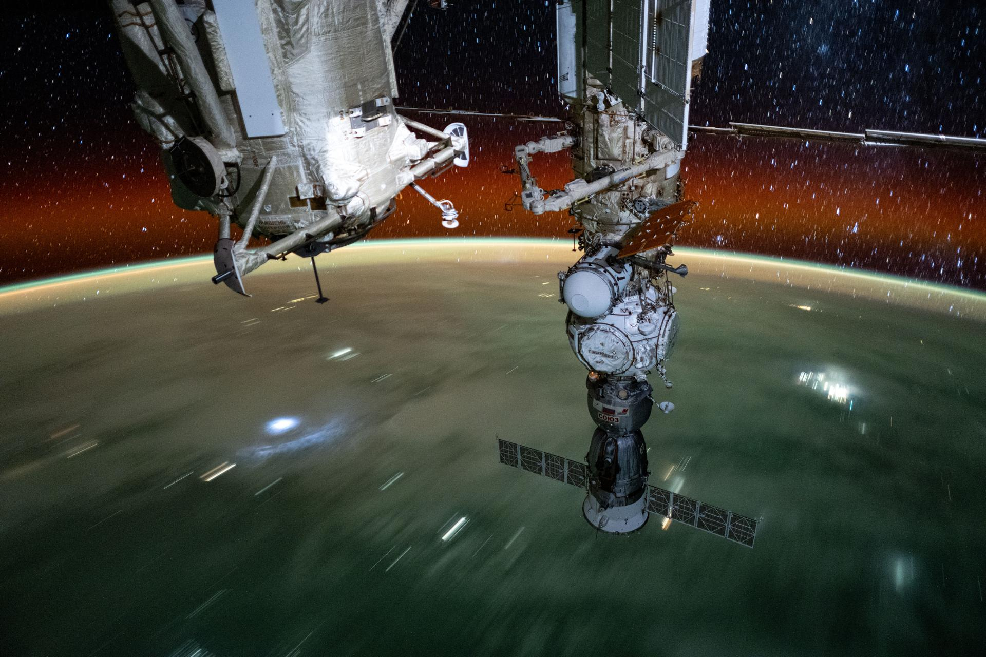 Two modules riddled with bars and joints and other mechanical nondescribables hang down from upward off frame. the right hangs lowest, with a black spacecraft docked at the bottom. The blurry curvature of earth rotates below as a horizon of stars streak above.