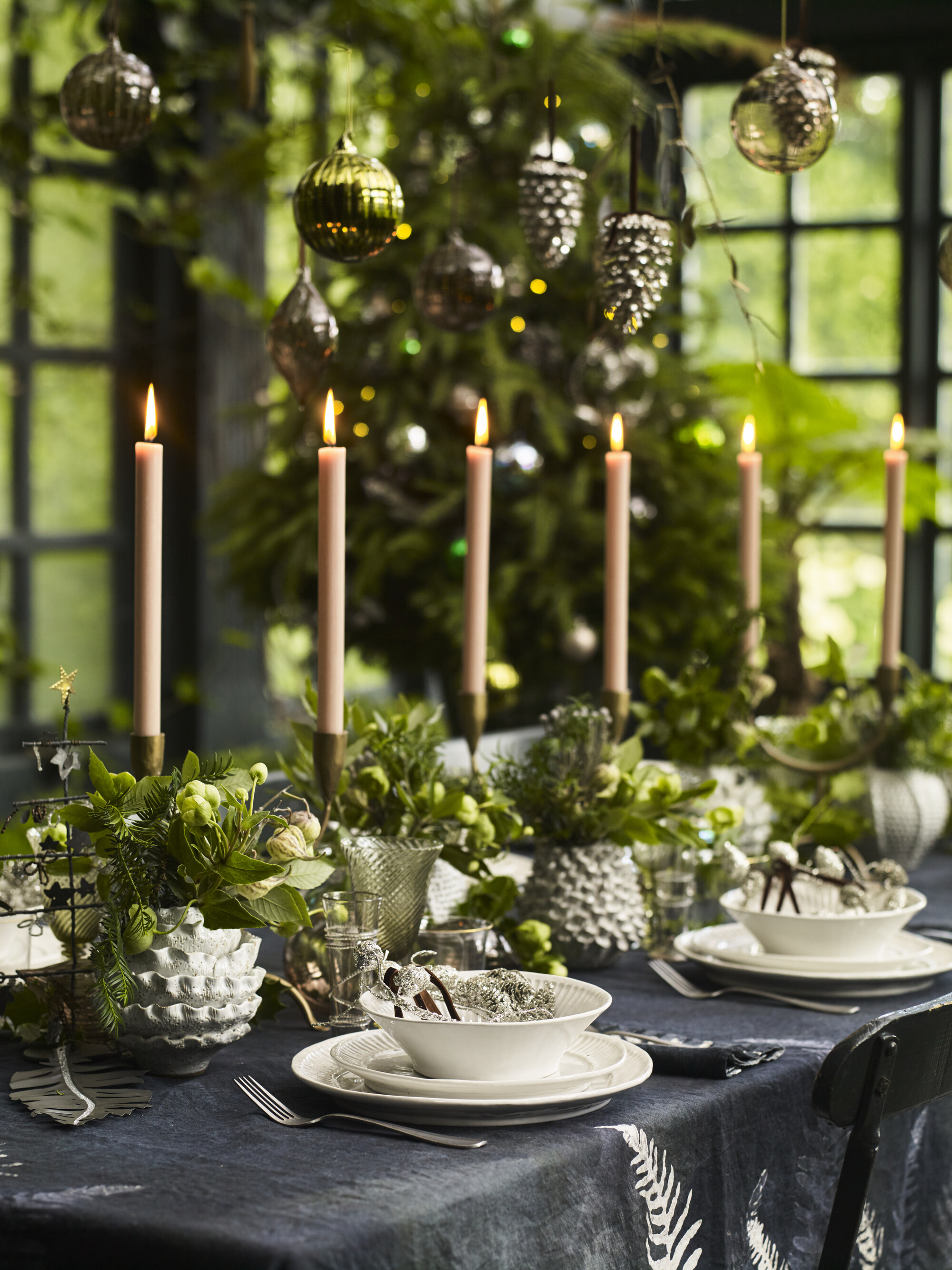 modern christmas table setting with black tablecloth, white dinnerware, brass candlesticks, fresh green foliage, ornaments hanging down from above