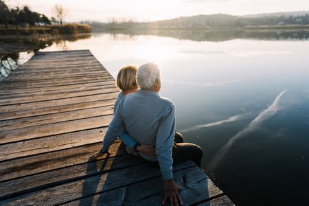 An older couple sits on a dock watching sunrise or sunset over a lake. She has her arm around him, and they face away from the camera.