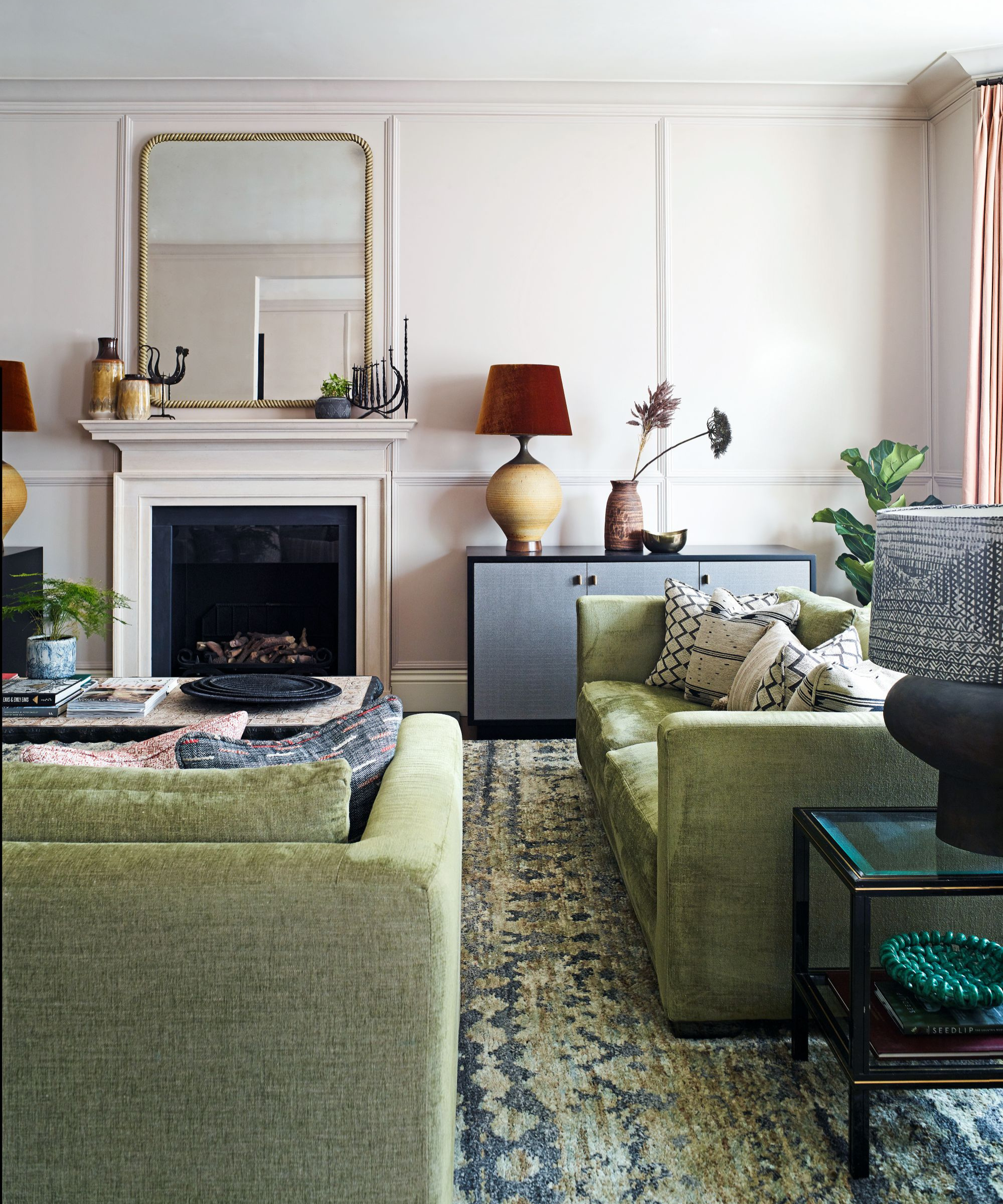 a living room with green sofas, a green and blue rug, pillows on the couches, and various lamps, vases, trays, and other miscellaneous decor