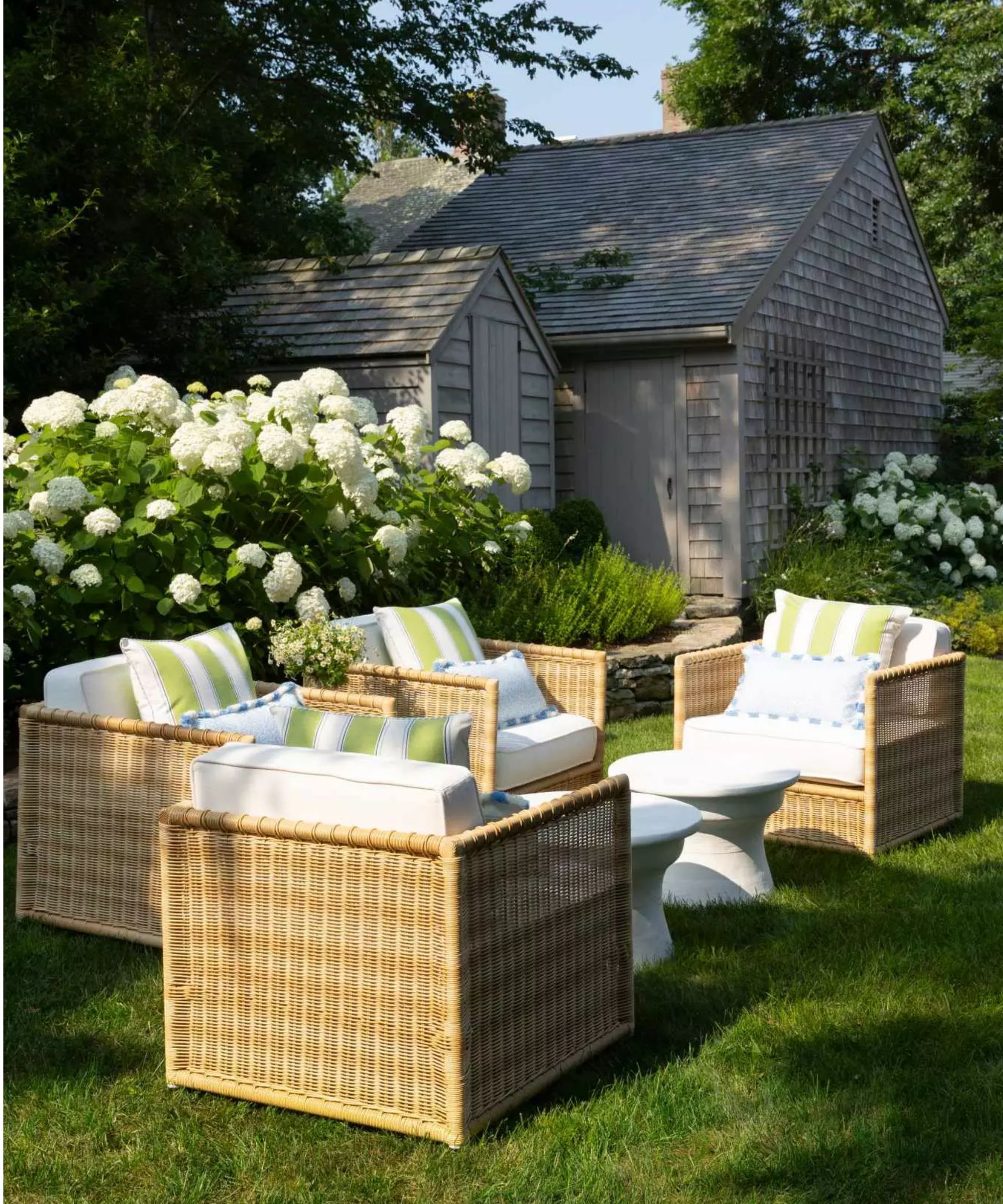 garden lawn with two outbuildings behind with large white hydrangea bushes and rattan patio furniture with white cushions and green and white striped throw pillows
