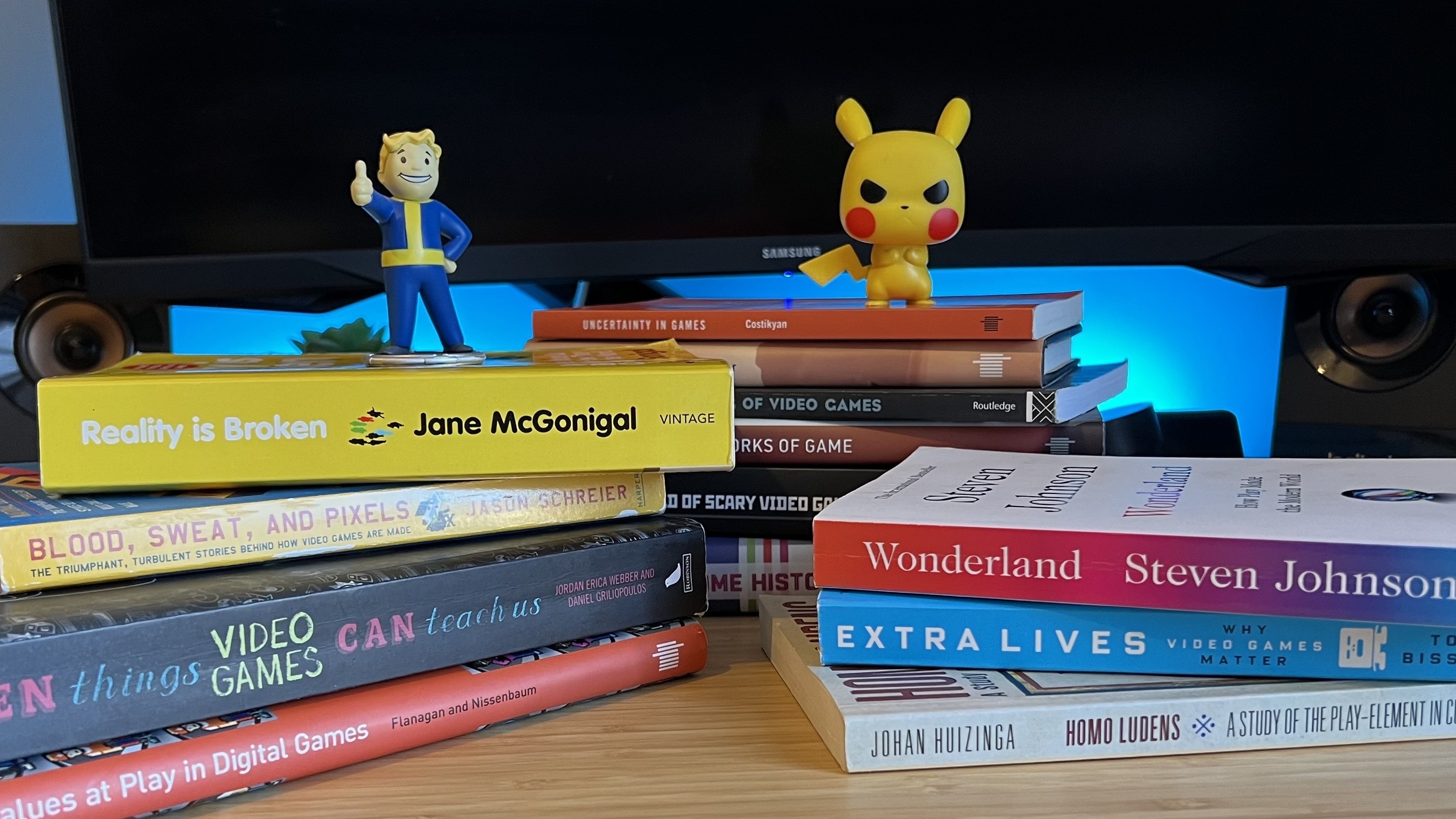 Gaming books stacked on a wooden desk with Fallout and Pokemon figures on top