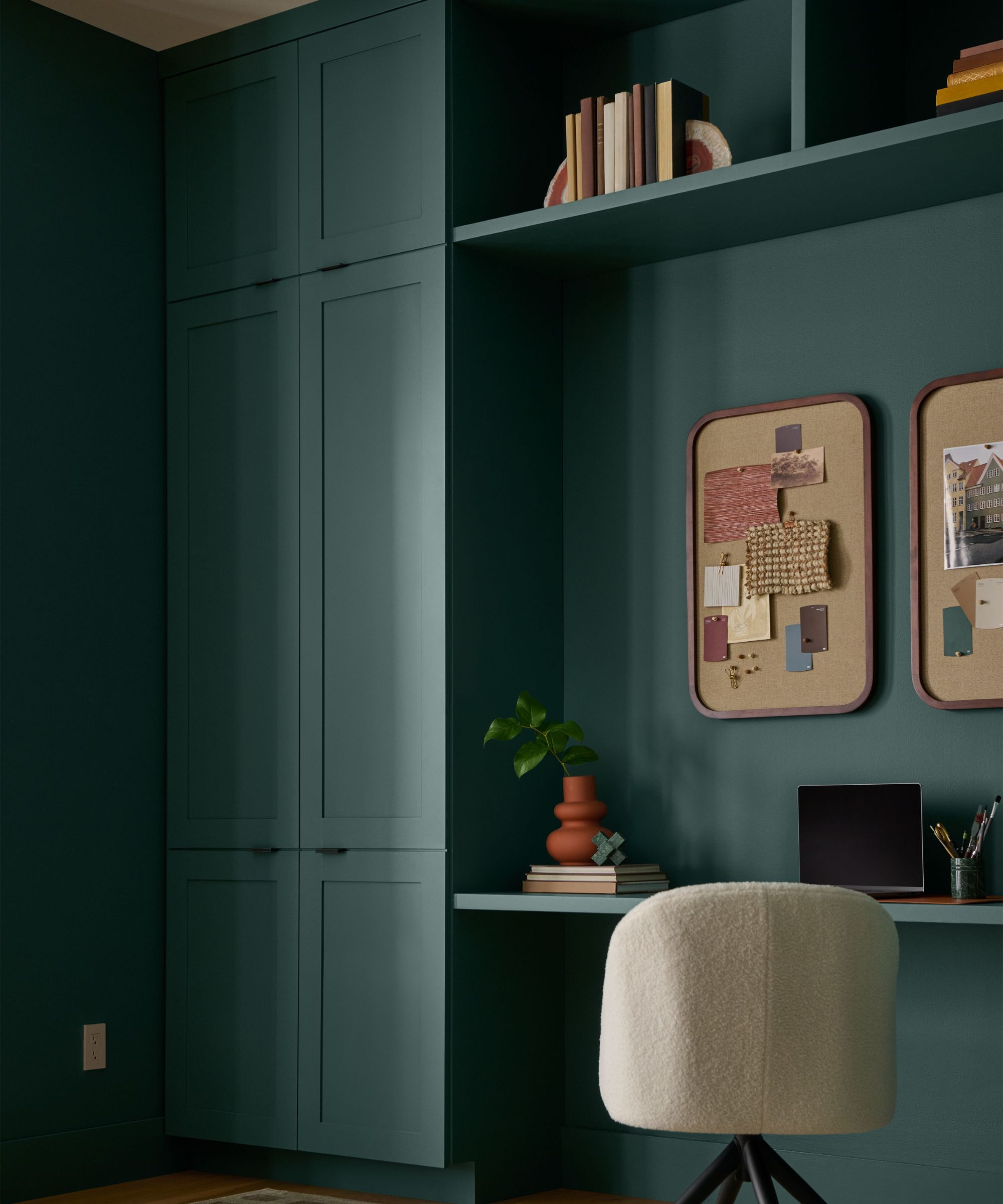 Home office painted in dark jade with built-in cabinetry and shelves, boucle desk chair, and pinboards