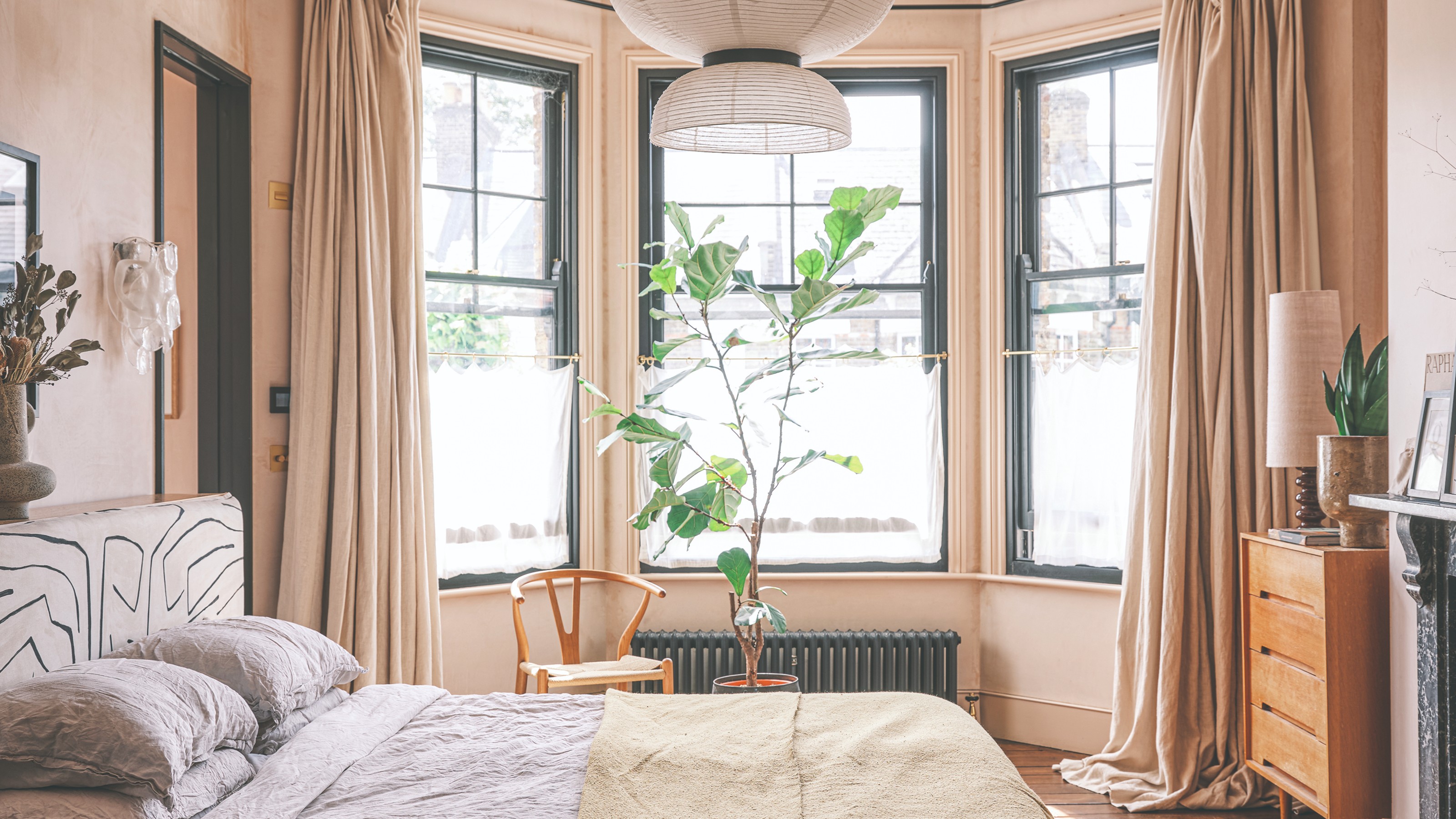 A bedroom painted in a soft, earthy pink with a bay window dressed with tonal floor-length curtains