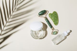A Jade roller along with a Jade Gua Sha tool, with facial serums, on a white background with the shadow of a leaf in view.