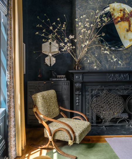 Corner of living room with dark blue wallpapered walls and fireplace, green and dark wood occasional chair, green and purple rug underneath, vase of flowers on top of mantle, yellow curtains