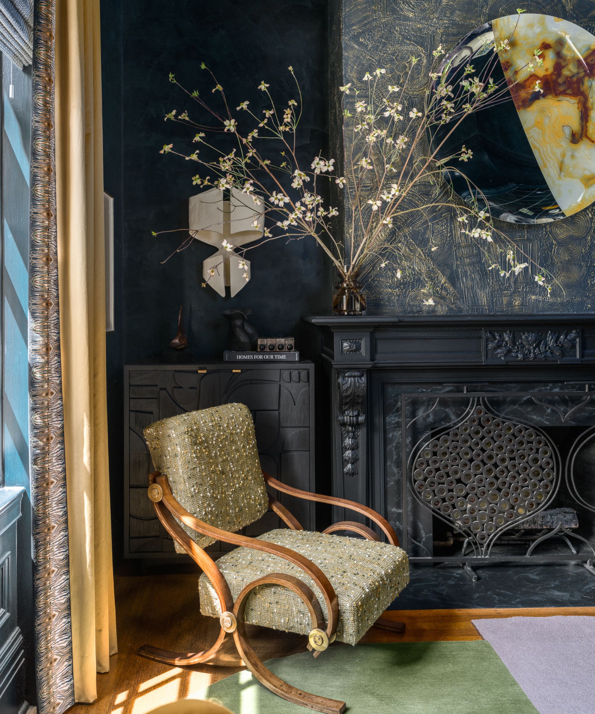 Corner of living room with dark blue wallpapered walls and fireplace, green and dark wood occasional chair, green and purple rug underneath, vase of flowers on top of mantle, yellow curtains
