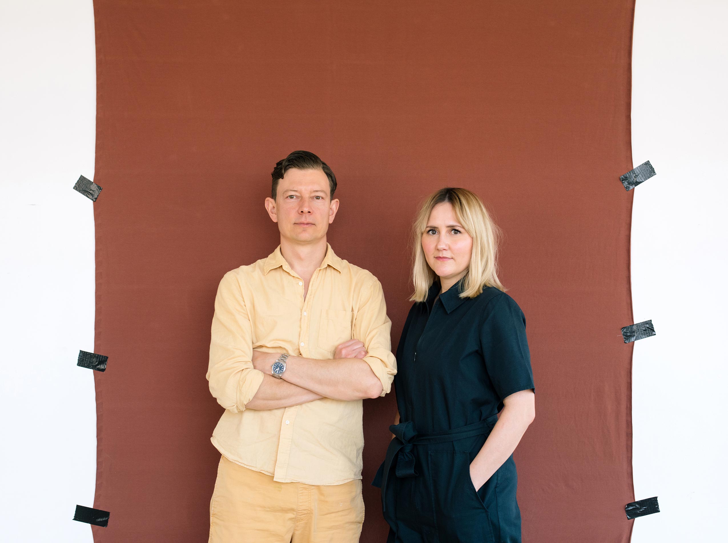 A man, dressed in pale yellow clothing, and a woman in a navy blue jumpsuit pose in front of a terracotta textile stitched to a white wall with tape.