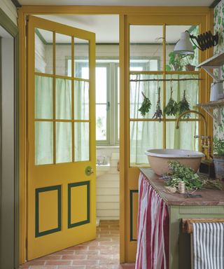 A kitchen with a wooden surface with a sink and lavender on top of it, and a bright yellow door opening up into a room with a white sink and light green framed windows