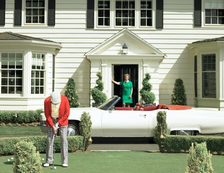 Retro photo of a man wearing outdated clothes golfing in front of his classic car and house. His wife looks on.