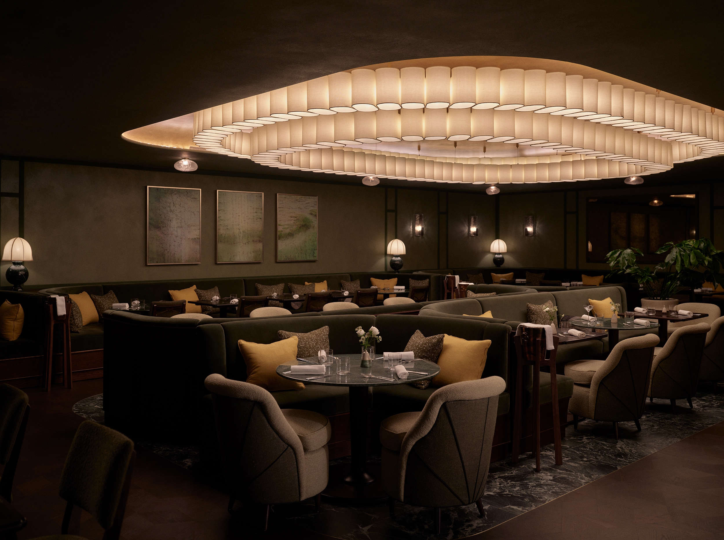 An atmospheric, luxurious restaurant features a multi-part, expansive chandelier made of lots of different lamp shades reunited over two concentric circular shapes, forest green furnishings, and mushroom-shaped table lamps.
