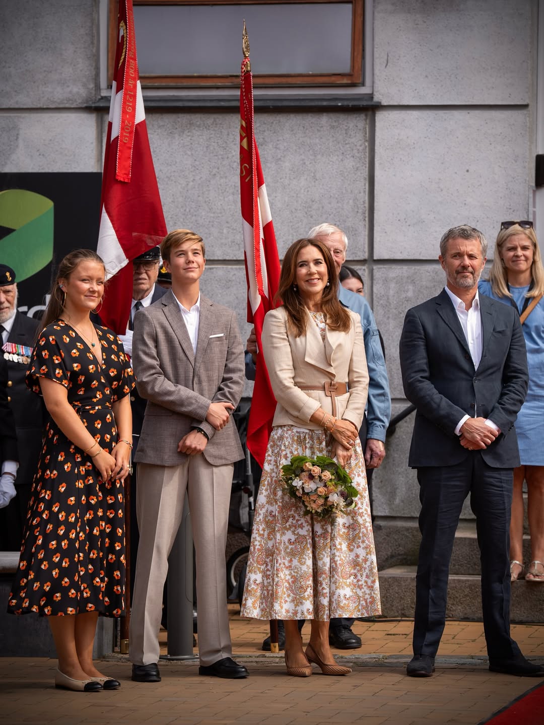 Princess Isabella, Prince Vincent, Queen Mary and King Frederik standing in a row in front of flags