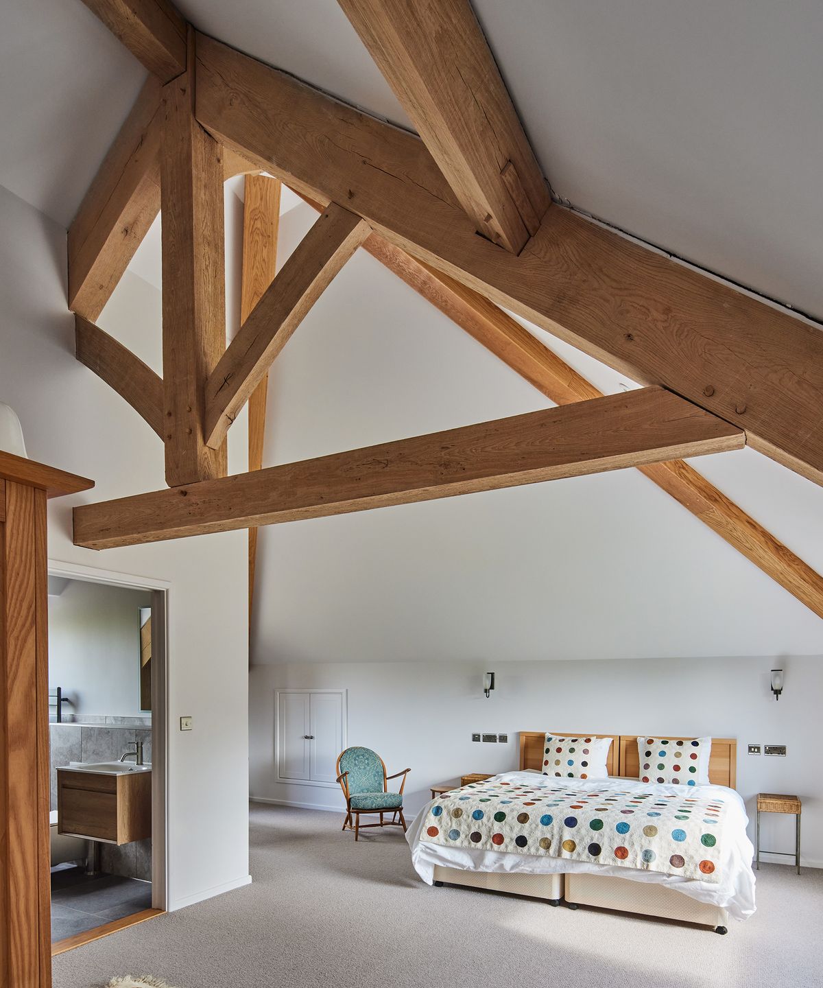 A large double bedroom with minimalist interiors. There are exposed beams in the ceiling which is high and pitched.