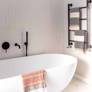 A modern bathroom with greige/taupe vertical bathroom wall tiles, a white ceramic bath tub, Terracotta throw, matt black heated towel rack and matt black shower accessories
