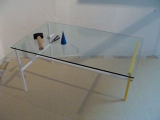 Three-legged glass side table photographed with a small hammer and 2 small cones (grey, white and blue) laid on it, against a white wall