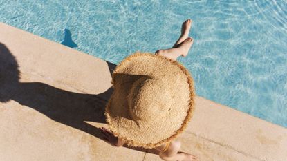 A pale girl with a large hat is sitting on the edge of a pool, possible wearing the best fake tan for pale skin