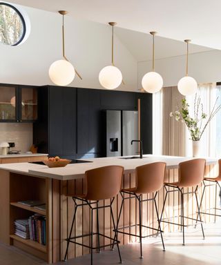 A kitchen with a concrete countertop kitchen island with a wooden base, three brown bar stool chairs in front of it, three globe gold pendant lights above it, and black cabinets in front of it