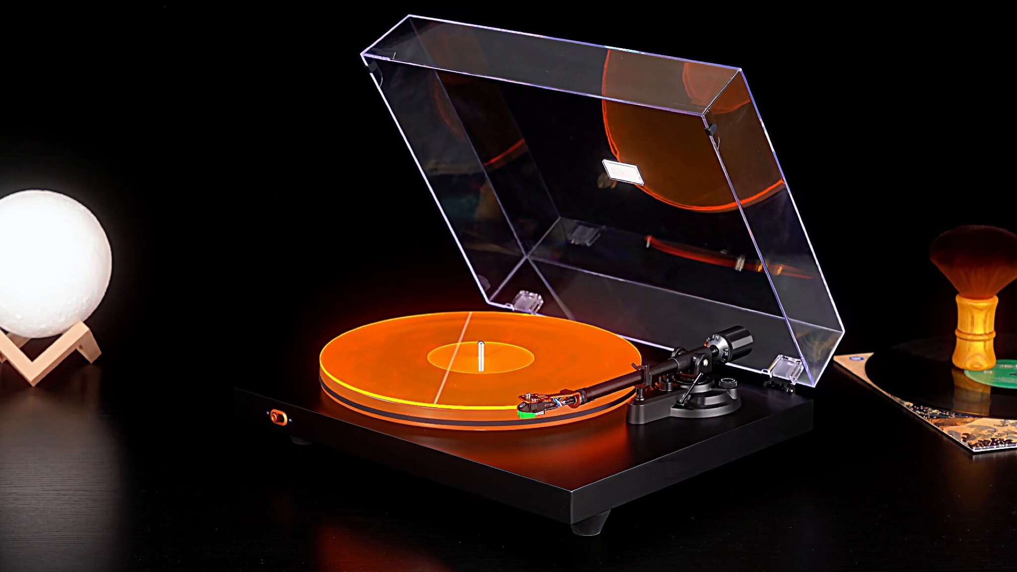 Angled view of the Fosi Audio 3 with its orange slip mat. The turntable is on a black surface with a lamp, a vinyl record and a brush beside it