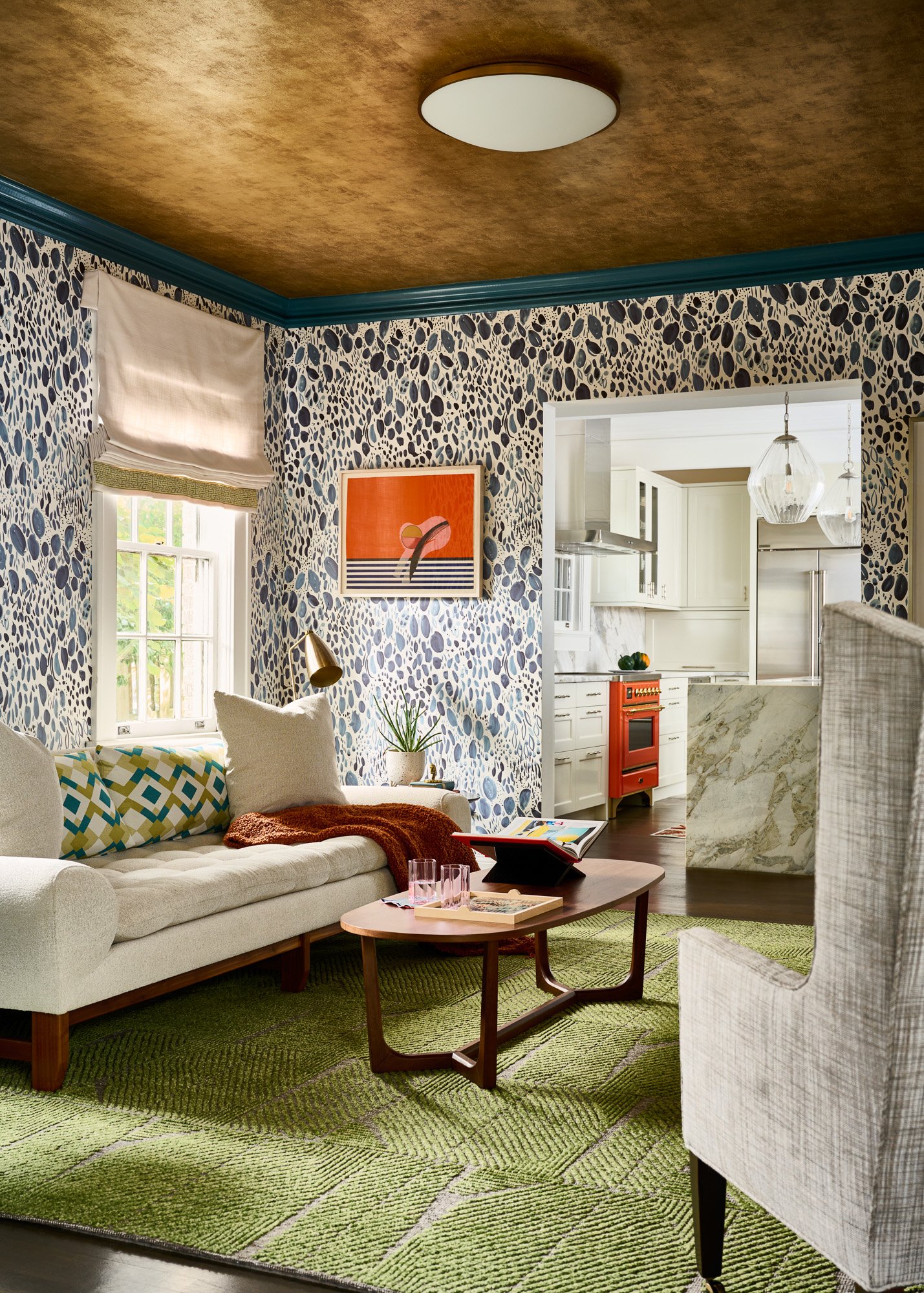 A whimsical living room with a green area rug, wallpapered walls, and teal trim. The furniture in the room is gray/cream and an orange piece of art sits on the wall.