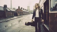 A young, AI-generated KK Downing steps off a bus holding a guitar case