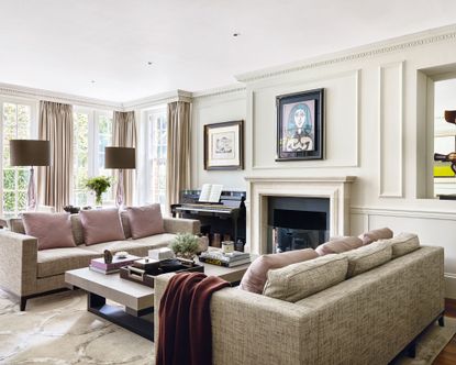 Bright living room with off white painted walls and paneling, spacious seating area with two gray three seater sofas, purple cushions, dark wood flooring with cream rug, twin gray table lamps, fireplace and piano 